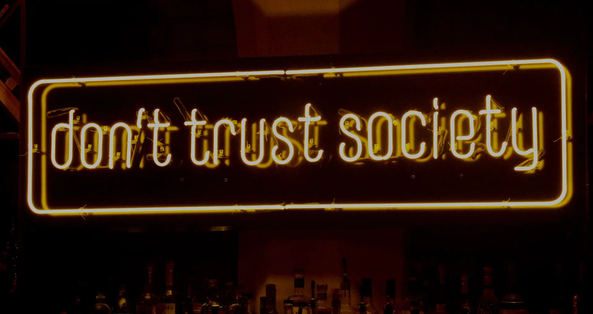 The 'don't trust society' neon sign glowing gold above the bar