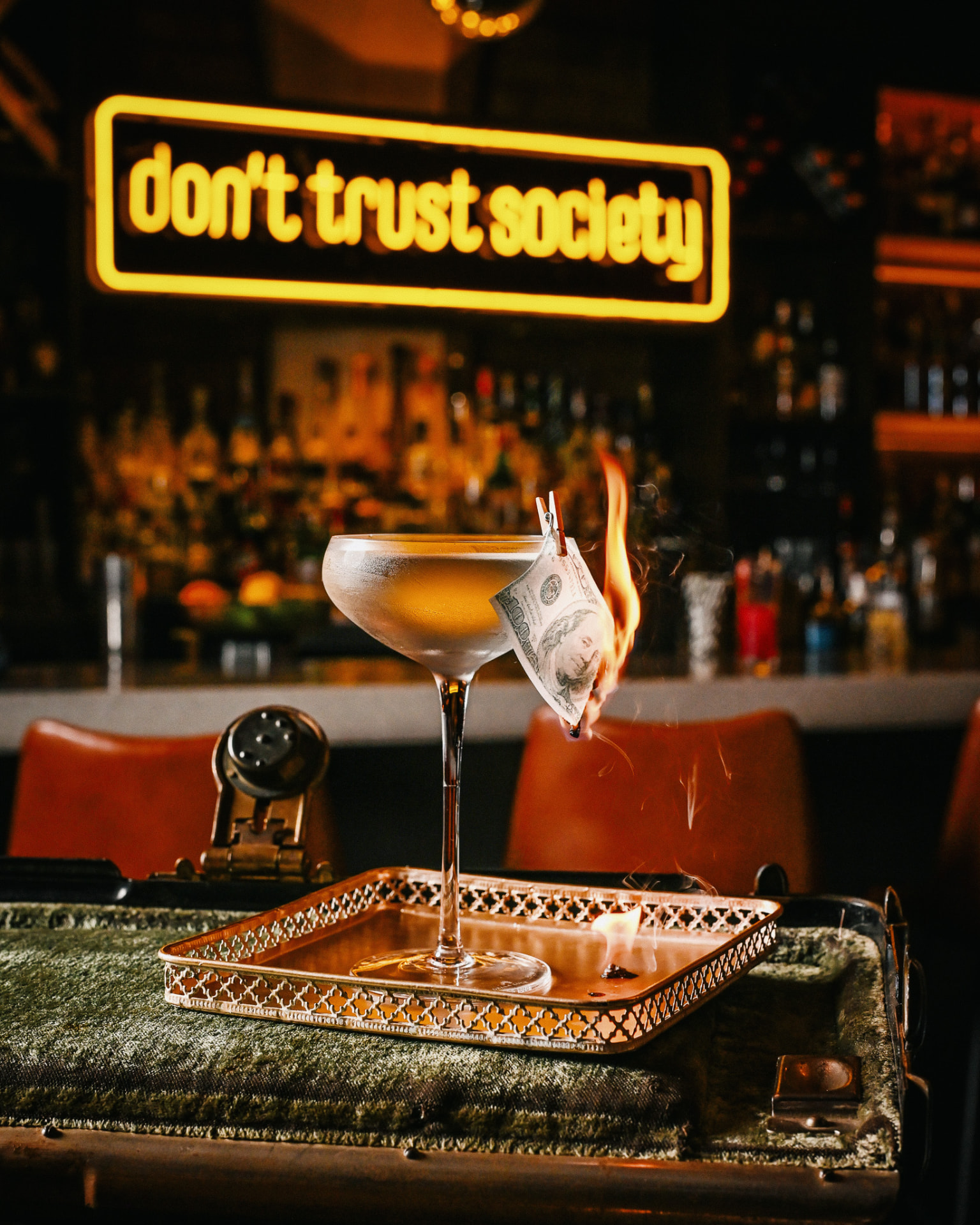 A flaming hundred-dollar-bill garnish on a coupe beneath the don't trust society neon sign